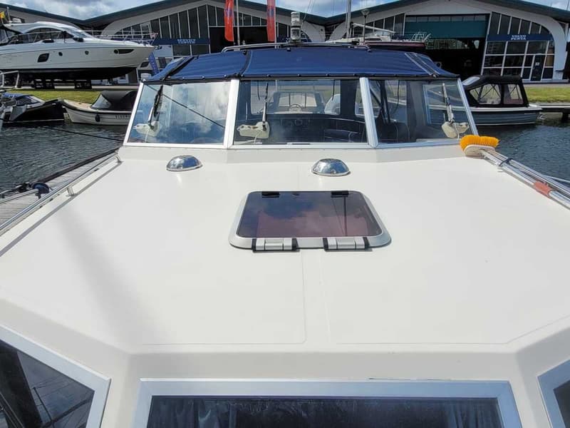 Een witte boot met een blauwe kap en ramen, liggend in het water bij een gebouw met de tekst "VOLVO PENTA". — Agder 840 OK