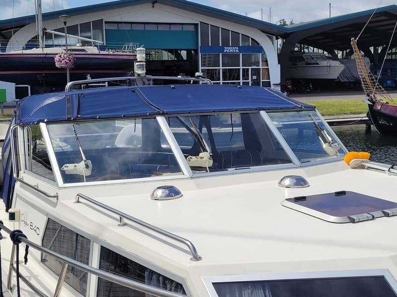 Een witte boot met een blauwe kap en voorruiten ligt in het water, met een gebouw met "Volvo Penta" op de achtergrond.