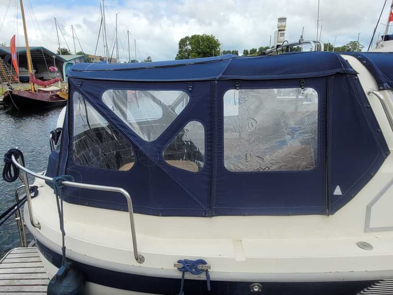 Een blauwe bootkap met doorzichtige ramen bedekt een deel van een boot, met een steiger en andere boten op de achtergrond.