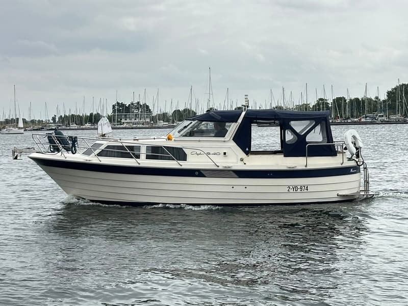 Een witte motorboot met blauwe accenten en een blauwe kap vaart op het water, met zeilboten op de achtergrond. — Agder 840 OK