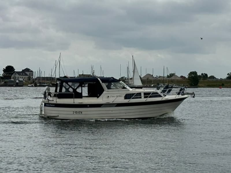 Een witte motorboot met blauwe kap vaart op het water, met een haven en huizen op de achtergrond onder een bewolkte lucht.