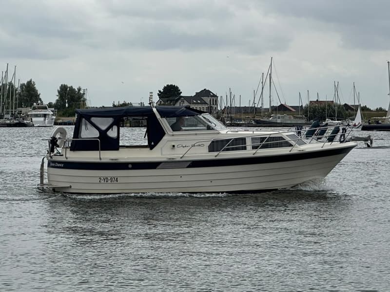 Een witte motorboot met blauwe accenten en een blauwe kap vaart op het water, met een haven op de achtergrond. — Agder 840 OK