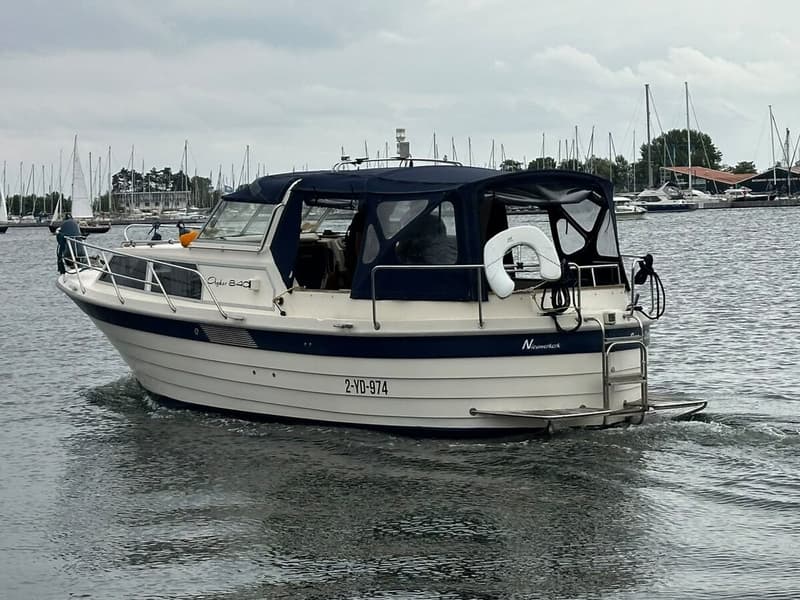 Een witte motorboot met blauwe accenten en een blauwe kap vaart op water met op de achtergrond andere boten. — Agder 840 OK