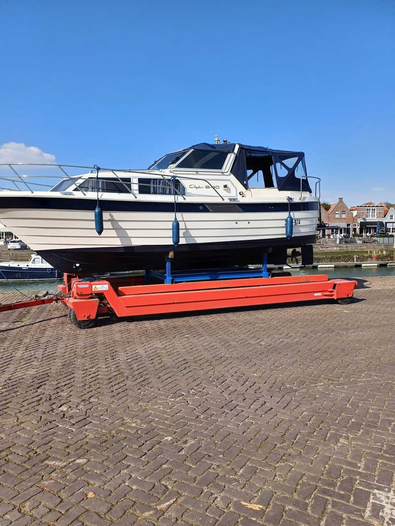 Een witte motorboot met blauwe accenten en een blauwe kap staat op een oranje trailer op een kade van klinkers.