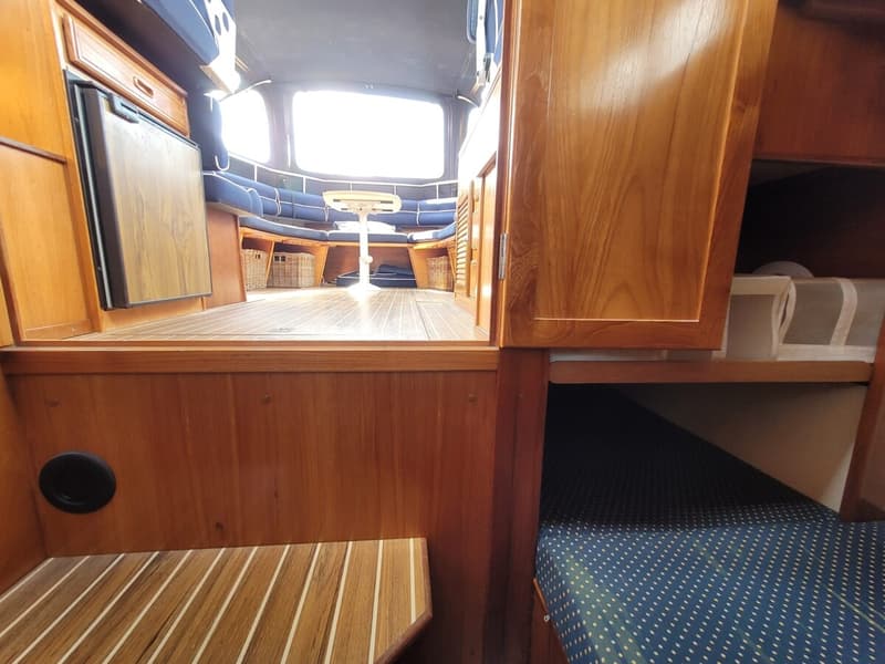 Een interieur van een boot met houten panelen, een koelkast, een zithoek met blauwe kussens en een bed. — Agder 840 OK