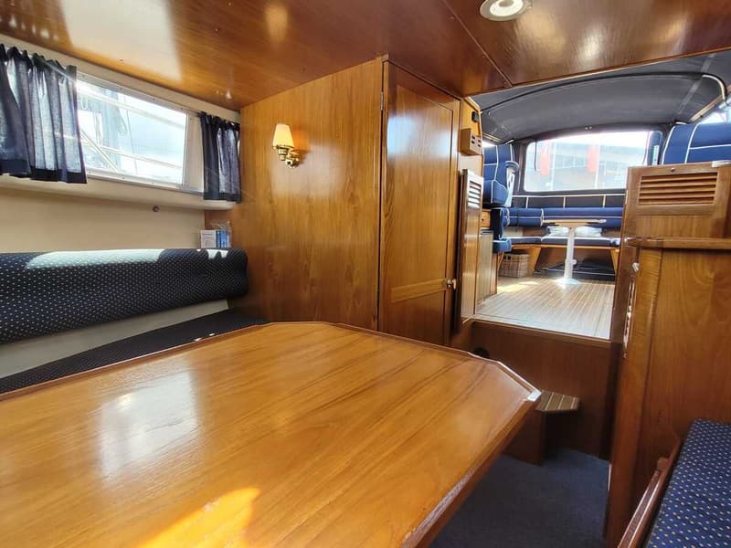 Een interieur van een boot met houten wanden en plafonds, blauwe kussens en een houten tafel op de voorgrond. — Agder 840 OK