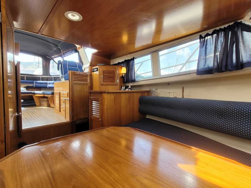 De foto toont het interieur van een boot met houten panelen, een tafel, banken met blauwe kussens en ramen met gordijnen.
