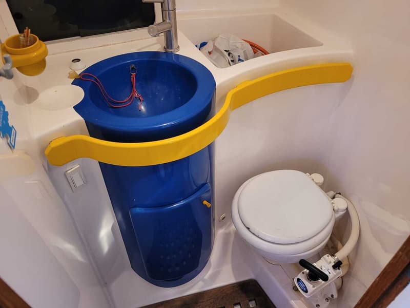 De foto toont een compacte toiletruimte met een blauwe wasbak, een kraan en een wit scheepstoilet.