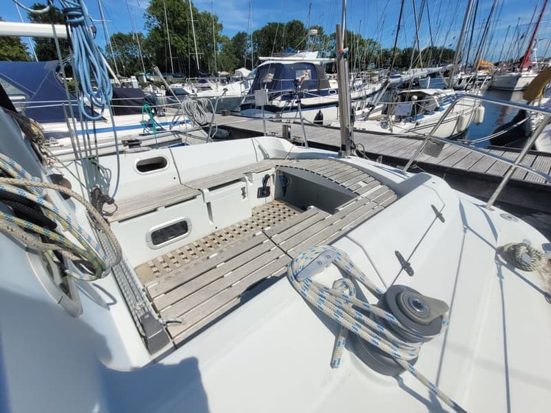 De foto toont de kuip van een witte zeilboot met houten vlonders en touwen, omringd door andere boten in een haven.
