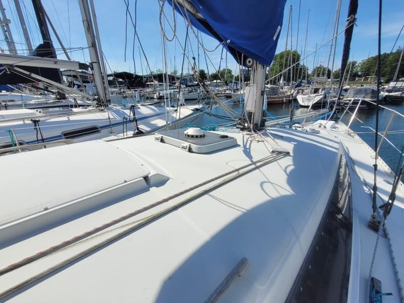 Een wit zeiljacht met een blauw zeil ligt in een jachthaven met andere boten onder een helderblauwe lucht.