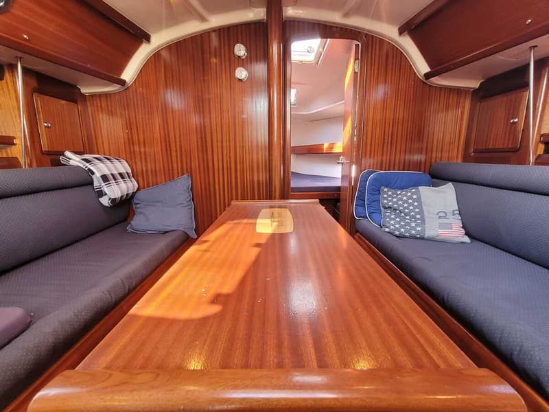 De foto toont het interieur van een boot met een houten tafel, twee banken met blauwe kussens en een doorgang naar een…