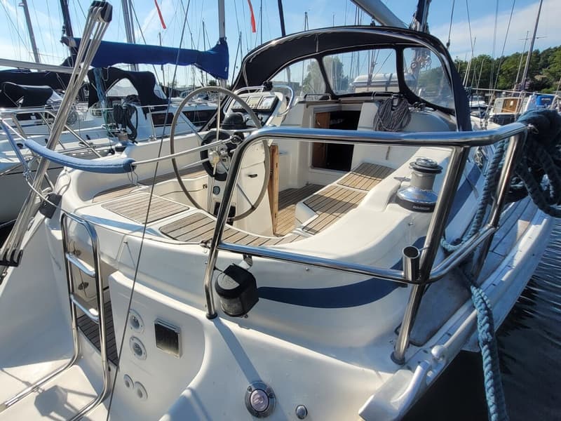 Een witte zeilboot met een blauwe streep, teakhouten dek en stuurwiel, ligt afgemeerd in een haven. — Bavaria 34-2