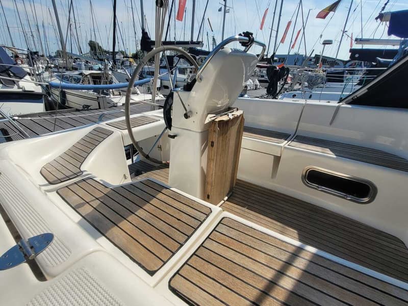 De foto toont de kuip van een zeilboot met een stuurwiel, houten vloerdelen en zitbanken, omringd door andere boten in een…