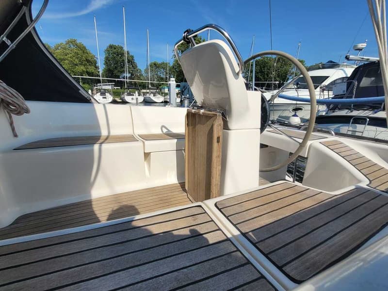De foto toont de kuip van een zeilboot met een stuurwiel, teakhouten vloer en zitbanken, met andere boten op de achtergrond.