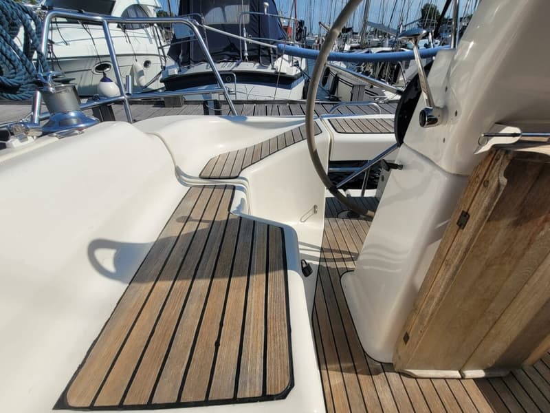 Een deel van een wit jacht met teakhouten dek en een stuurwiel, liggend in een haven met andere boten. — Bavaria 34-2