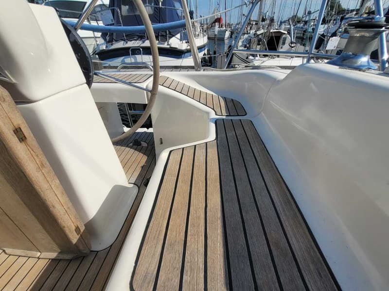 Een detailfoto van het dek van een zeilboot met houten latten en een wit stuurwiel, met andere boten op de achtergrond.