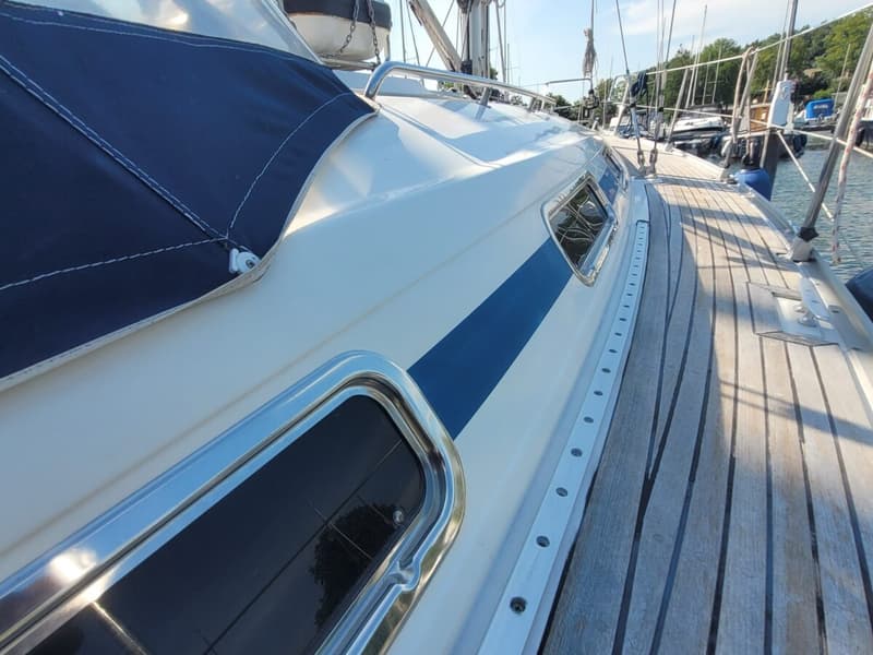 De foto toont de zijkant van een witte zeilboot met een blauwe streep, patrijspoorten en een houten dek. — Bavaria 34-2