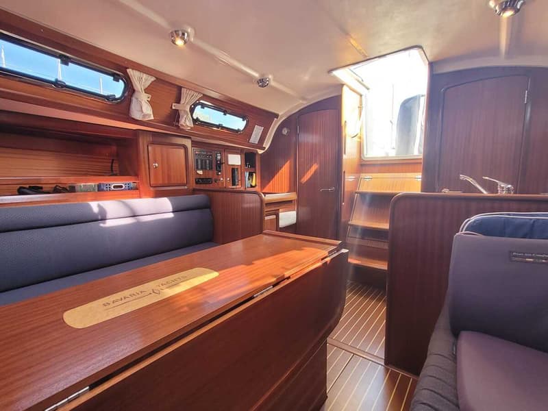 De foto toont het interieur van een boot met houten afwerking, een tafel met "BAVARIA YACHTS" erop, en blauwe banken.