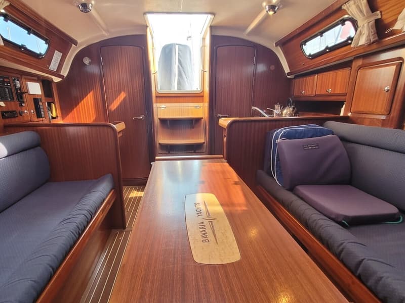 De foto toont het interieur van een boot met houten afwerking, blauwe kussens en een tafel met het Bavaria Yachts logo.