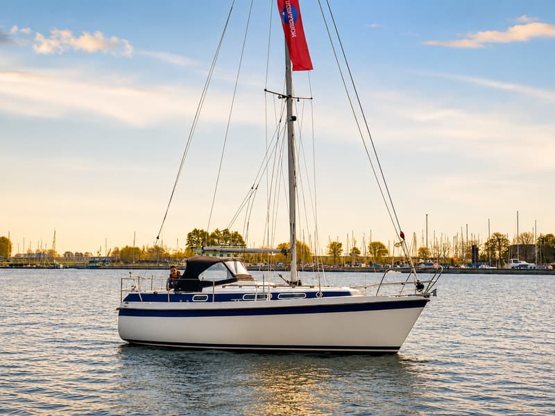 Een witte zeilboot met een blauwe streep op de romp ligt op het water, met een rode vlag in de mast en een haven op de…