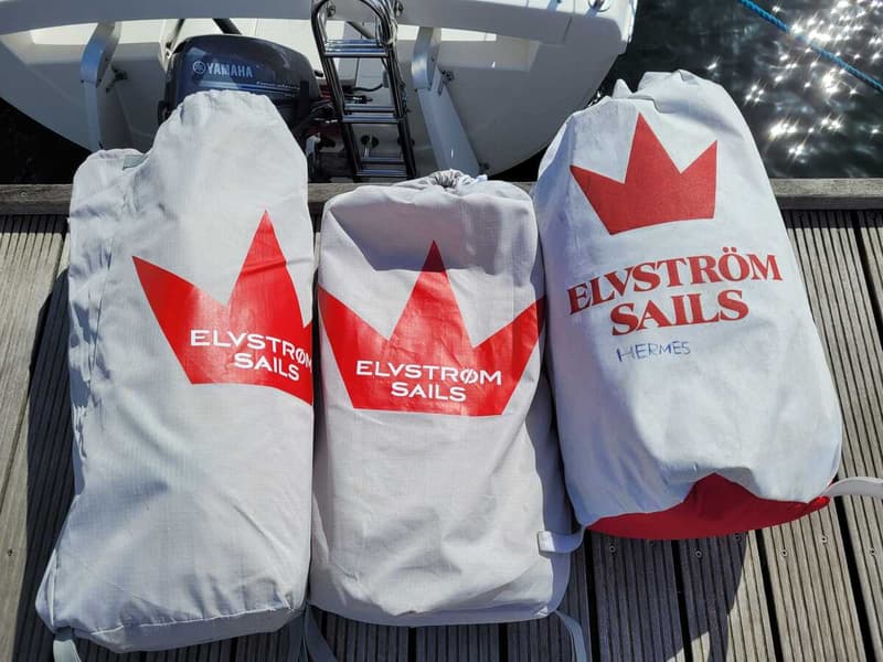 Drie grijze zeilzakken met rode opdruk "ELVSTRÖM SAILS" en een kroonlogo liggen op een houten steiger.