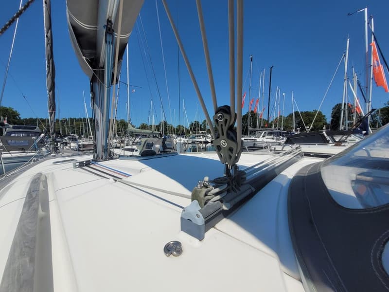 Een dek van een zeilboot met een mast, zeil, blokken en lijnen, en andere boten in de achtergrond onder een blauwe lucht.