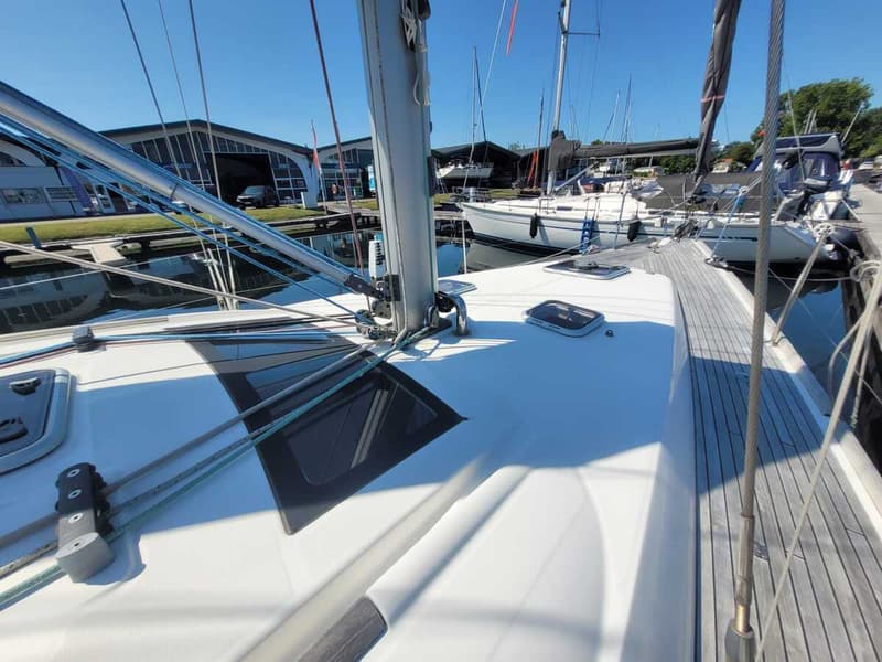 Een witte zeilboot met een houten dek en een mast staat in een haven met andere boten en gebouwen op de achtergrond.