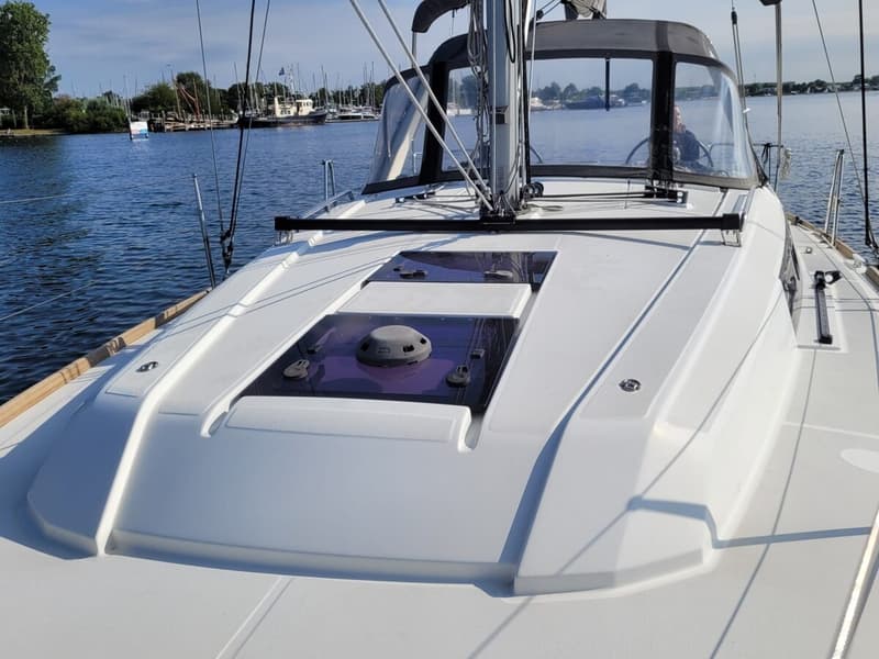 Een witte zeilboot met een donkere buiskap en ramen vaart op een waterweg met andere boten en bomen op de achtergrond.