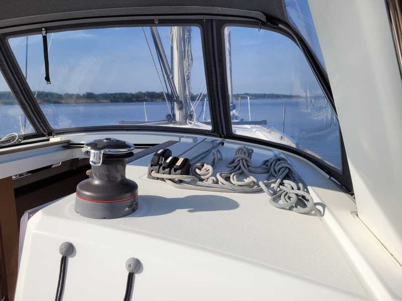 Een lier en touwen op het dek van een zeilboot, met uitzicht op water en een beboste oever. — Beneteau Oceanis 35