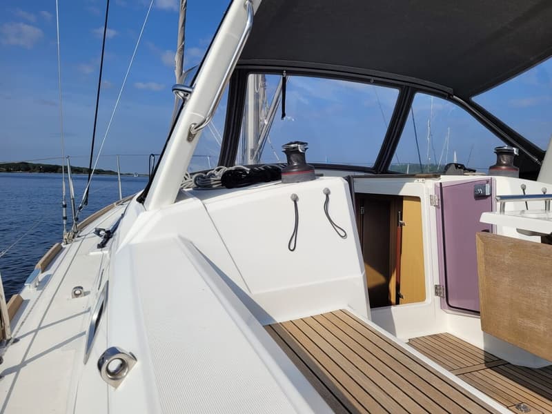 Een deel van een witte zeilboot met een teakdek, een open luik en een lier, met water en land op de achtergrond.