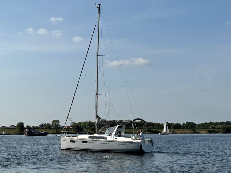 Een witte zeilboot met een opgerold grootzeil en een persoon aan het roer vaart op een waterweg. — Beneteau Oceanis 35