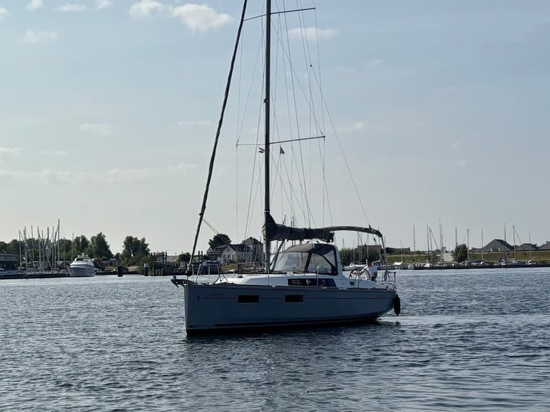 Een zeilboot met een grijze romp en een donkergrijze buiskap vaart op water met een jachthaven op de achtergrond.