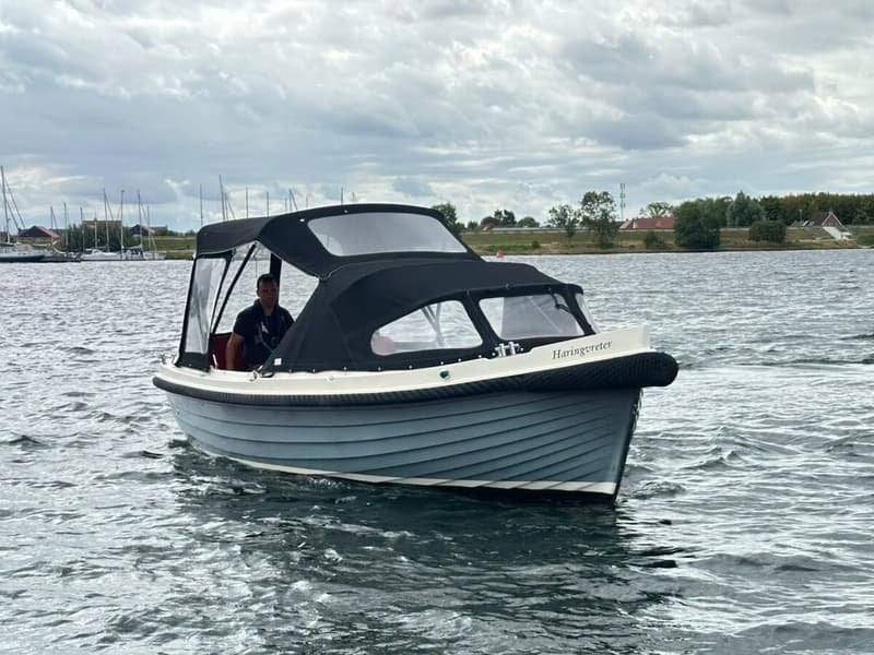 Een blauwe sloep met een zwarte buiskap en een man aan het roer vaart op water met een bewolkte lucht. — Interboar 22