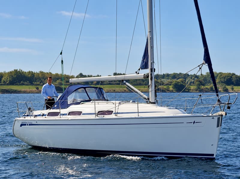 Een witte zeilboot met blauwe strepen en een blauwe buiskap vaart op het water, met een man in een blauw overhemd aan boord.