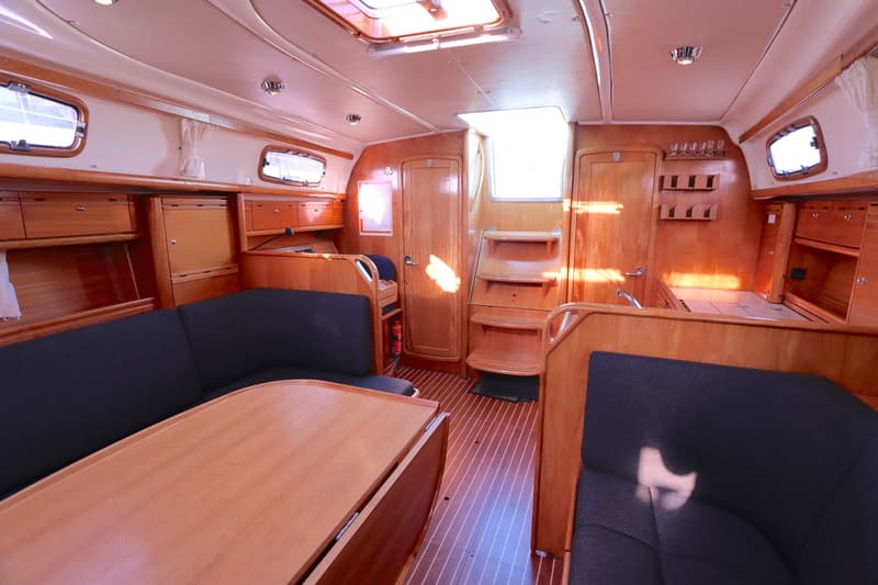 De foto toont het interieur van een boot met houten afwerking, donkerblauwe kussens, een tafel en ramen.