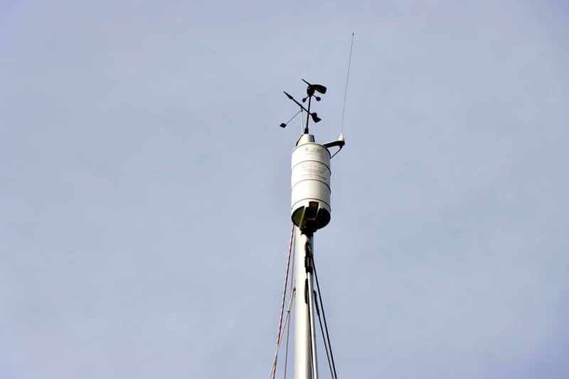 Een witte mast met een windvaan, windmeter en antenne tegen een lichtgrijze lucht.