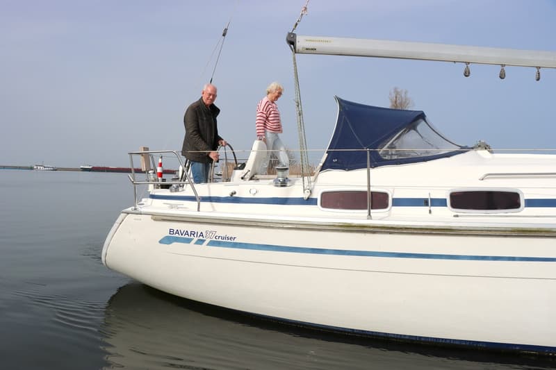 Een witte zeilboot met blauwe strepen en de tekst "BAVARIA 37 cruiser" vaart op het water met twee mensen aan boord.