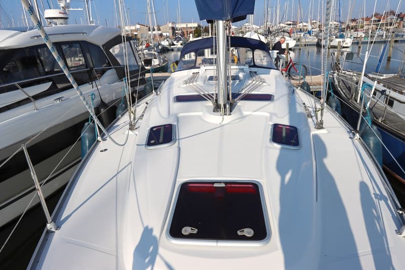 Een bovenaanzicht van het dek van een witte zeilboot met luiken en een mast, omringd door andere boten in een haven.