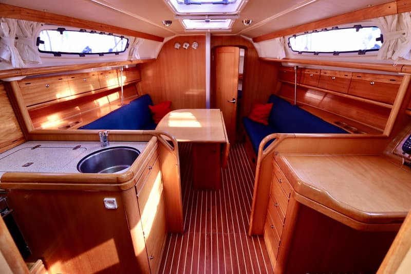De foto toont het interieur van een boot met houten afwerking, blauwe banken, een tafel en een kombuis met spoelbak.