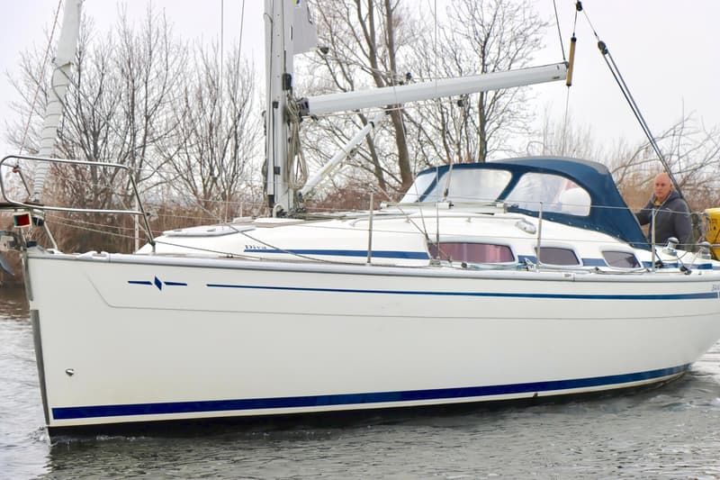 Een witte zeilboot met blauwe strepen en de naam "Diva" op de boeg, met een man in een donkere jas aan boord. — Bavaria 31