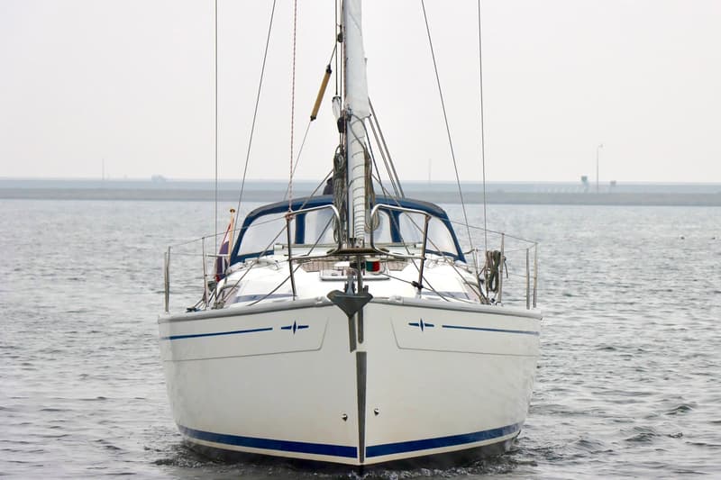 Een witte zeilboot met blauwe accenten en een opgerold zeil staat in het water, met een dijk op de achtergrond. — Bavaria 31