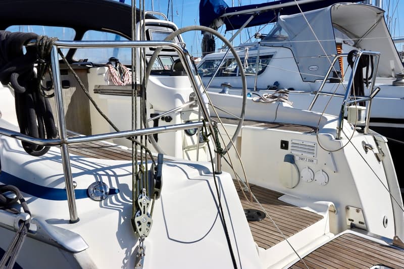 Een deel van een witte zeilboot met een stuurwiel, reling en touwen, met een andere boot op de achtergrond. — Bavaria 31