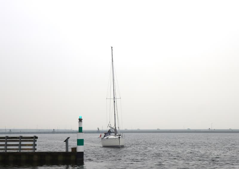 Een witte zeilboot vaart langs een groen-witte vuurtoren en een houten steiger op een grijze dag. — Bavaria 31