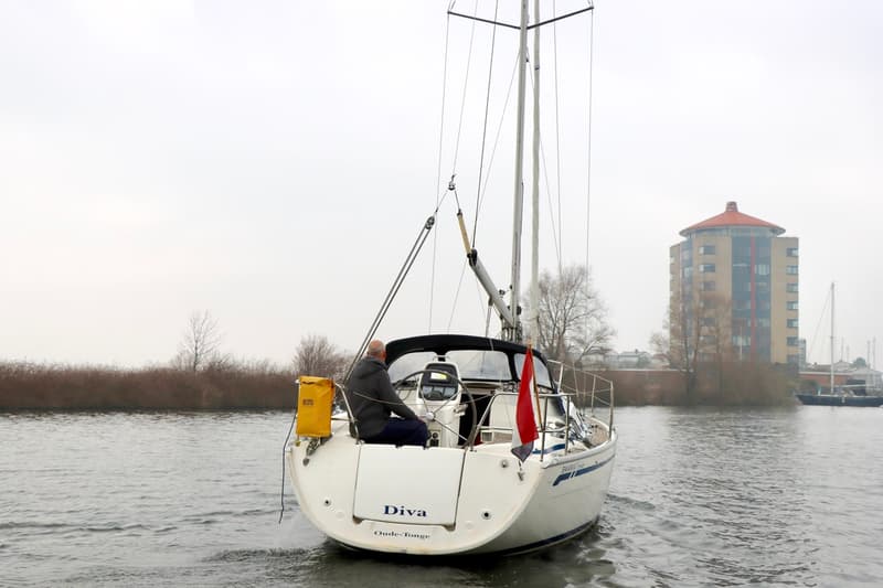 Een zeilboot met een man aan het roer vaart op een waterweg, met een gebouw op de achtergrond. — Bavaria 31