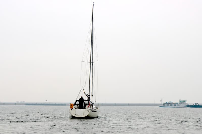 Een witte zeilboot met een persoon aan boord vaart op water, met een ander schip en een dijk op de achtergrond. — Bavaria 31