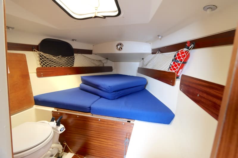 De foto toont het interieur van een bootkajuit met blauwe kussens, een toilet, een brandblusser en opbergnetten.
