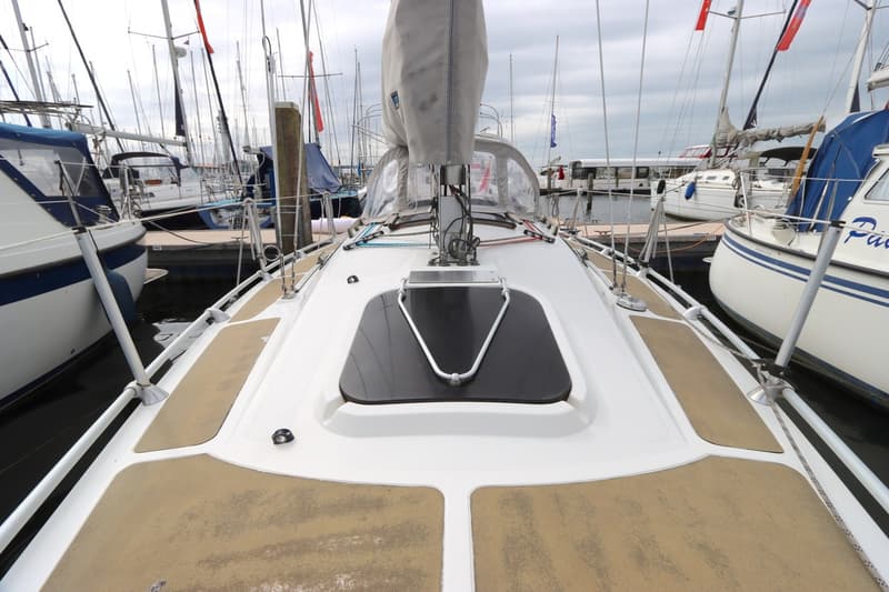 Een bovenaanzicht van het dek van een zeilboot, met een luik en een opgerold zeil, omringd door andere boten in een haven.