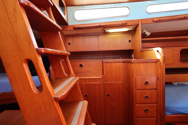 Een houten kajuitinterieur met een trap, kasten, lades, patrijspoorten en een bed met blauw matras.