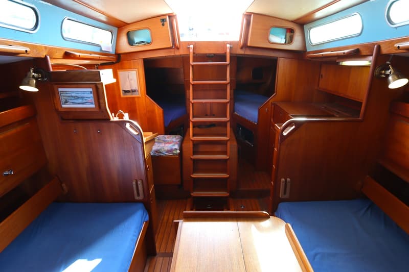 De foto toont het interieur van een boot met houten afwerking, blauwe kussens op de bedden en een ladder in het midden.
