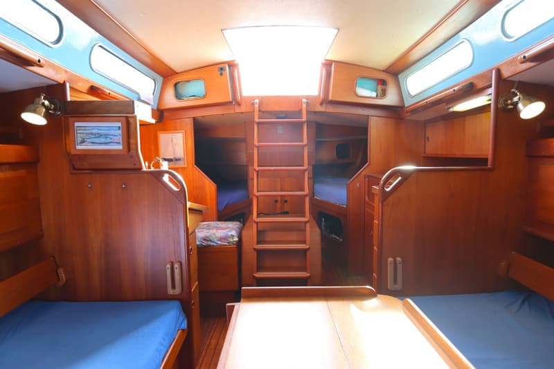 De foto toont het interieur van een boot, met houten wanden, blauwe kussens op bedden, een ladder en een dakraam.